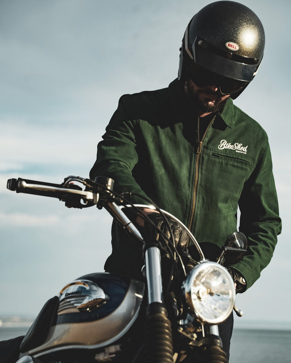 Model wearing the Bike Shed Moto Co. Twill Green logo jacket standing next to a motorcycle.