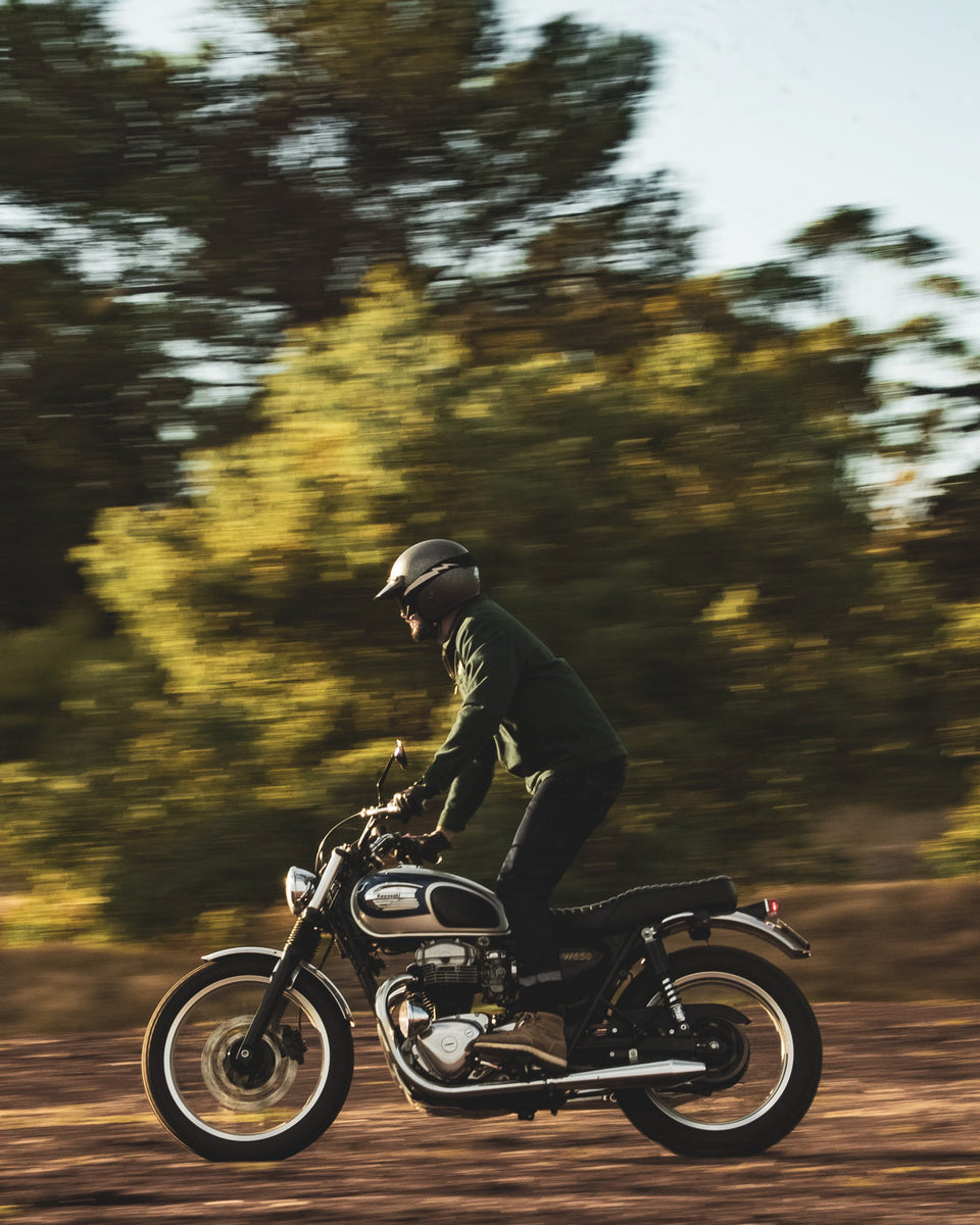 Model wearing the Bike Shed Moto Co. Twill Green logo jacket standing riding a motorcycle off road on a dirt path.