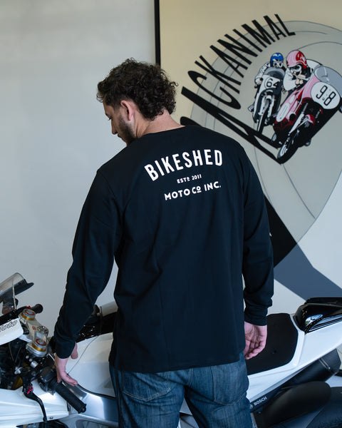 Model wearing Bike Shed Moto Co ESTD Long sleeve t-shirt in black next to a motorcycle in front of art work