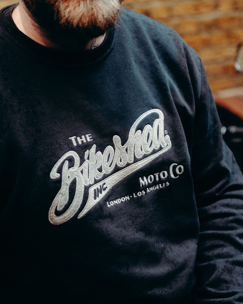 Model wearing Bike Shed Moto Co Black Applique Sweatshirt next to a motorcycle on a brick driveway