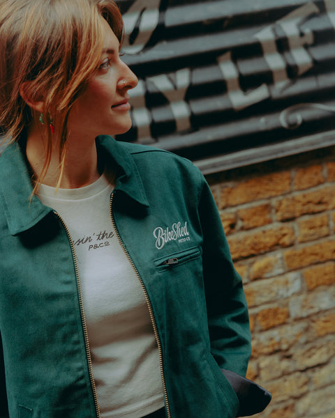 Model wearing green motorcycle jacket in front of a brick wall with graffiti.